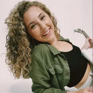 With a joyful smile and curly hair framing their face, the person proudly holds a guitar. They're dressed in a black top layered with a green shirt, standing against an unsin plain background.
